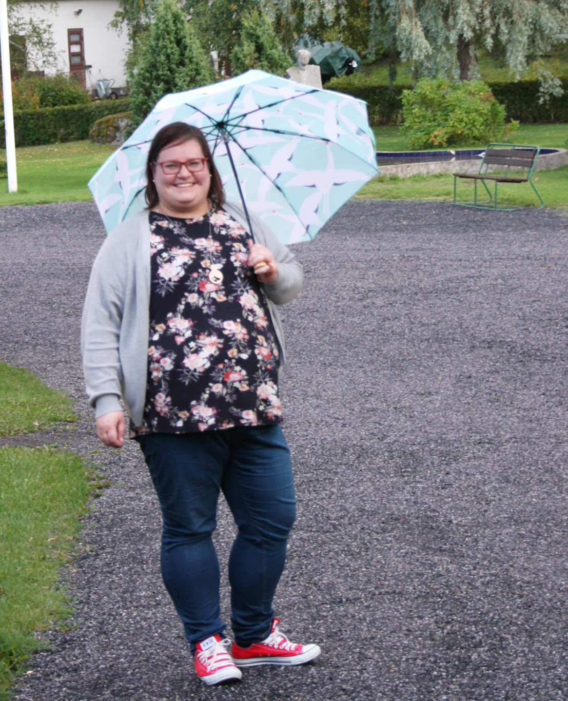 hymyilevä silmälasipäinen nainen farkuissa ja kukkapaidassa sinisävyisen sateenvarjon kanssa soraisella pih...
