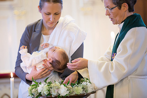 naispuolinen pappi kastaa äidin sylissä olevaa vauvaa
