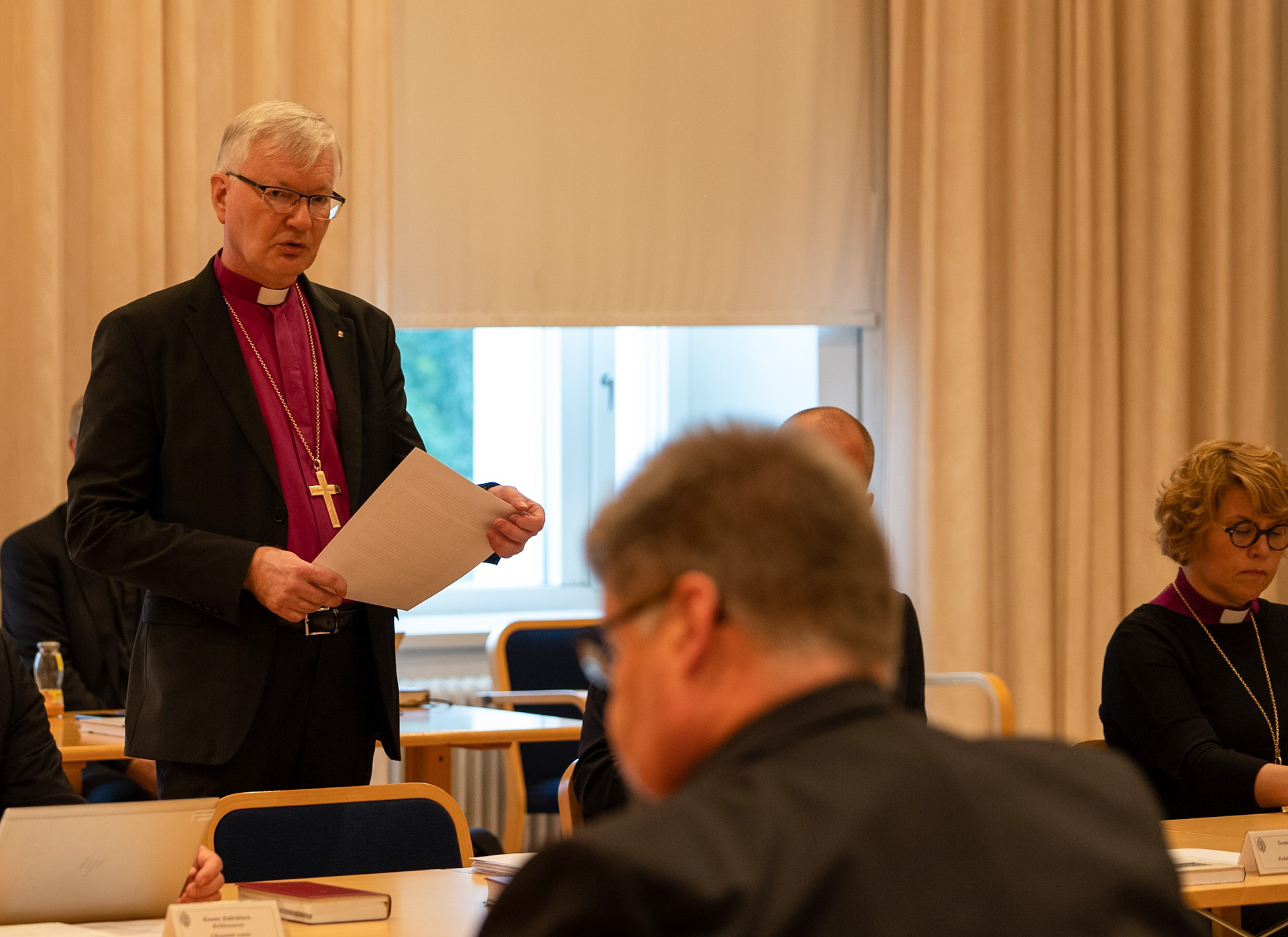 silmälasipäinen mies piispan asussa puhuu paperi  kädessä kokouksessa